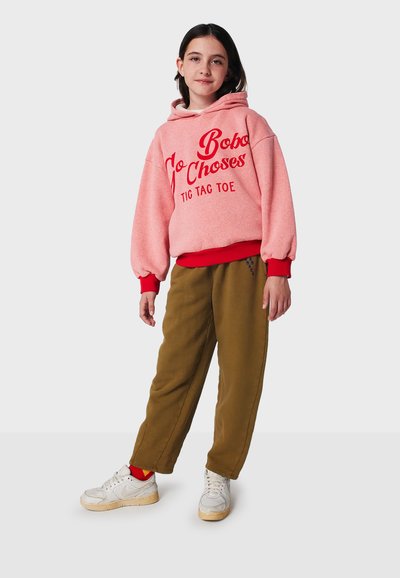 Rosa Fleece-Hoodie mit rotem Grafiktext, kombiniert mit khakifarbenen Trainingshosen mit seitlicher Stickerei und weißen Sneakers mit roten Akzenten.