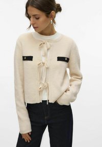 Crème kleurige cardigan met zwarte accentzakken, gestrikt aan de voorkant, en een zachte textuur. Lange mouwen en een cropped ontwerp.
