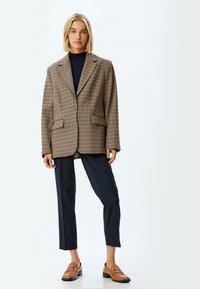 Karierte Blazer in Braun und Beige mit breitem Revers, zwei Fronttaschen und Zwei-Tasten-Verschluss, kombiniert mit dunklen Hosen und braunen Slippern.