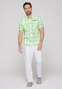 Camisa de cuadros verdes de manga corta con dos bolsillos en el pecho y botones a presión, combinada con pantalones de lino blancos y chanclas azules.
