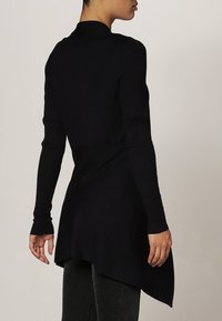 Svart ribbad långärmad topp med hög krage och asymmetrisk fåll, med en texturerad design och åtsittande ärmar.