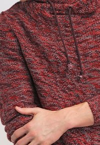 Roter und grauer strukturierter Strickpullover mit einem Schalkragen und verstellbaren Kordeln, der einen lockeren Schnitt und umgeschlagene Ärmel hat.