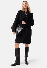 Cappotto di lana nero con ampi rever, abbinato a una maglietta a righe bianco e nero, borsa nera con motivo coccodrillo e stivali neri al ginocchio.