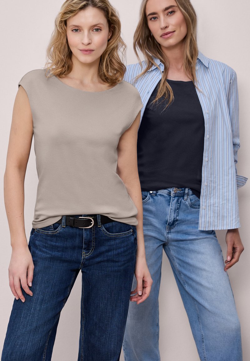 Deux femmes posant en tenues décontractées : l'une porte un haut sans manches beige avec un jean foncé, l'autre un haut noir, une chemise à rayures et un jean clair.
