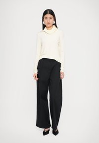 Crème col roulé top gecombineerd met stijlvolle zwarte high-waisted wijduitlopende broeken en puntige zwarte hakken, op een effen witte achtergrond.