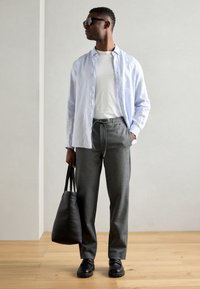 Cămașă bleu deschis cu guler, purtată peste un tricou alb, pantaloni gri, pantofi negri și o geantă de umăr neagră, pe un fundal neutru.