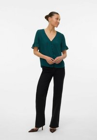 Blusa turchese con scollo a V, maniche corte arricciate e chiusura frontale con bottoni, abbinata a pantaloni larghi neri e tacchi a punta marroni.