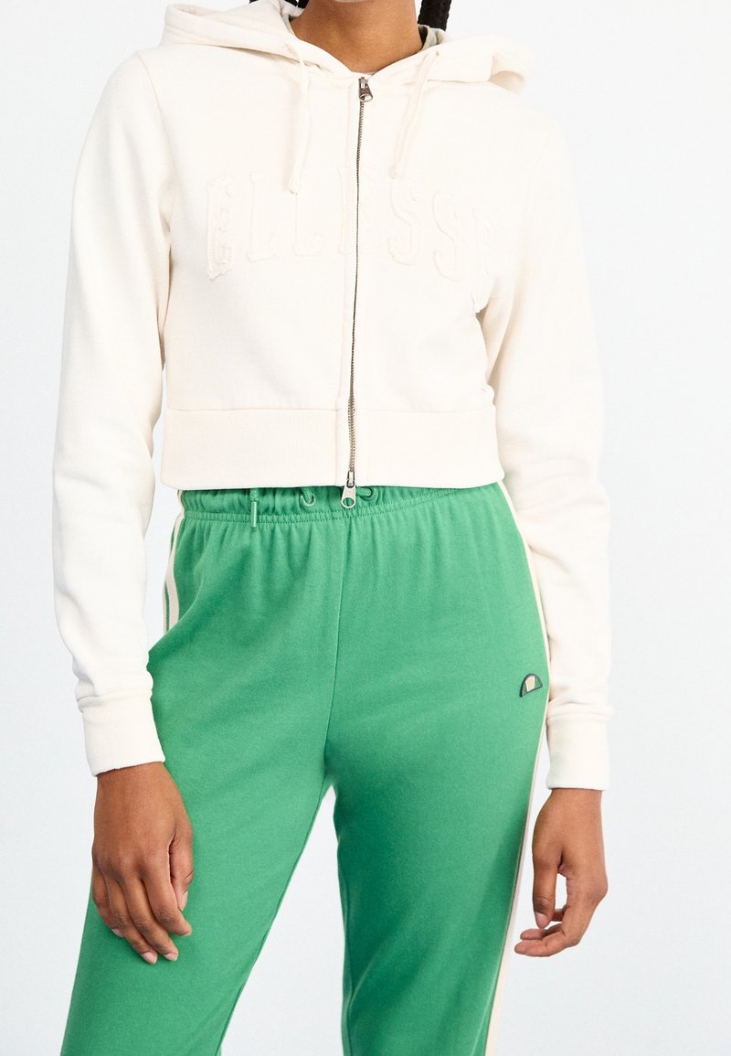 Felpa beige corta con zip frontale e scritta in rilievo, abbinata a pantaloni della tuta verdi con vita elastica e strisce laterali.