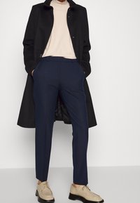 Abrigo negro, top beige, pantalones azul marino de corte ajustado y zapatos beige con suelas negras. El abrigo cuenta con un cierre de botón.