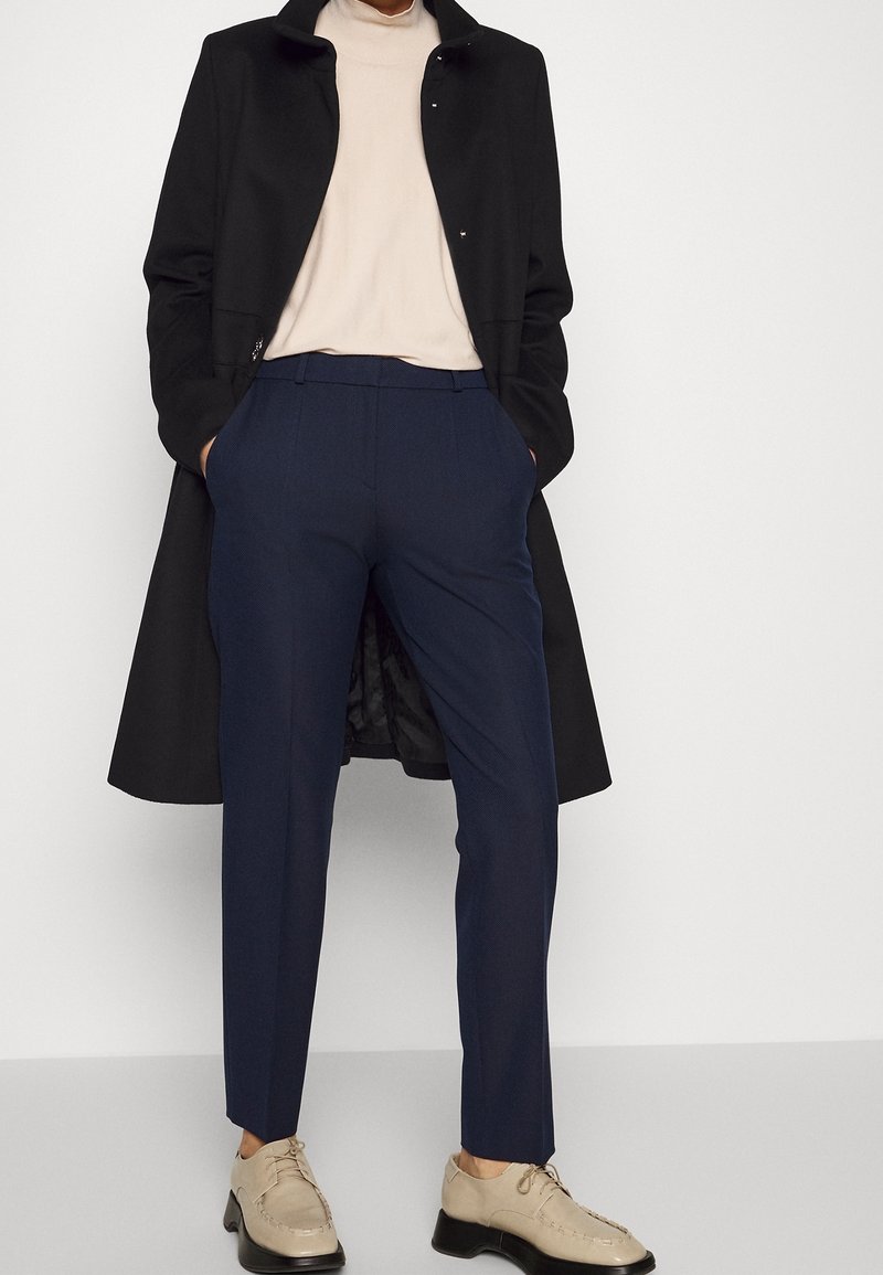 Abrigo negro, top beige, pantalones azul marino de corte ajustado y zapatos beige con suelas negras. El abrigo cuenta con un cierre de botón.