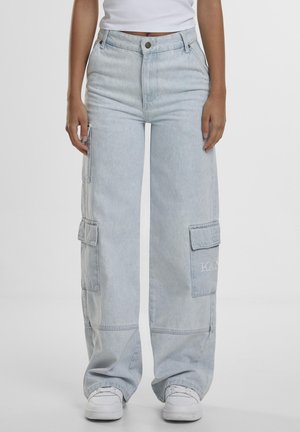 Persoană purtând blugi cargo largi de culoare albastru deschis cu buzunare cu clapetă și pantofi sport albi, stând în picioare pe un fundal simplu.