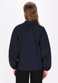Camicia blu navy con colletto alto, dettaglio sul retro arricciato e maniche ampie a sbuffo. Tessuto liscio con un leggero lucido, design minimalista.