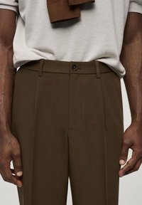 Braune, maßgeschneiderte Hosen mit glatter Textur, einzelnen Vorderfalten und Knopfverschluss, kombiniert mit einem hellgrauen Oberteil und braunem Strickaccessoire.