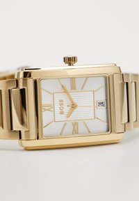 Montre rectangulaire en or avec un cadran blanc, des chiffres romains et un motif texturé. Elle comprend un affichage de la date et un bracelet métallique.