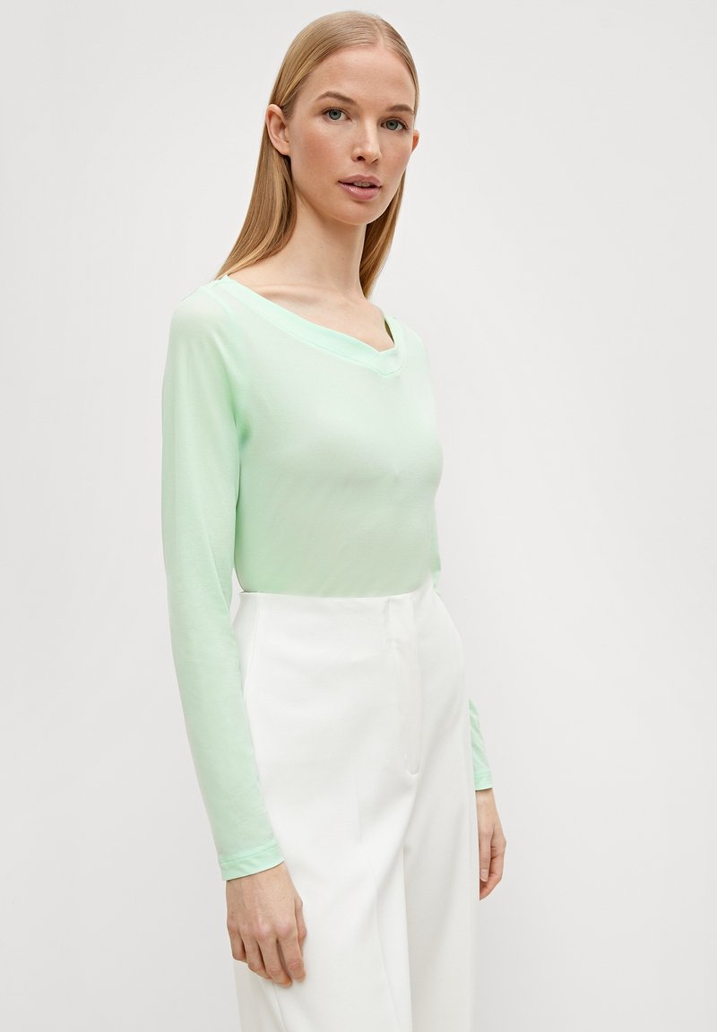 Lichtgroen langemouwen shirt met een brede V-hals, gemaakt van zachte stof. Gecombineerd met hoge witte broeken.