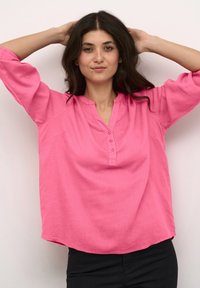 Chemise longue à manches longues rose clair avec un col en V et trois boutons, présentant une coupe décontractée et une texture de tissu douce.