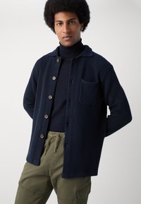Marineblau strukturierte Strickjacke mit Knopfverschluss, einer einzelnen Brusttasche und einem lässigen Kragen, getragen über einem marineblauen Rollkragenpullover und olivgrünen Hosen.