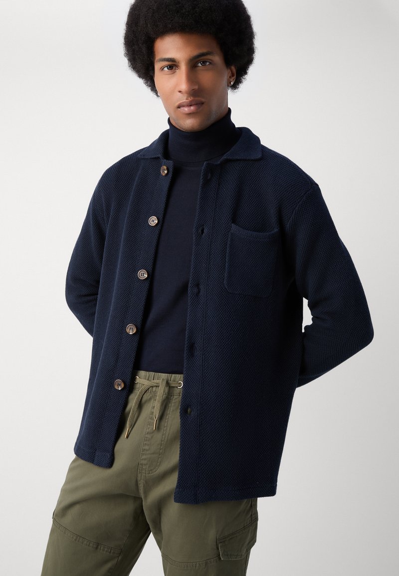 Cardigan en maille bleu marine avec des boutons sur le devant, une poche poitrine simple et un col décontracté, porté sur un col roulé bleu marine et un pantalon olive.