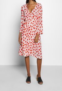 Frau in einem weißen Midikleid mit roten Herzmustern und langen Ärmeln, kombiniert mit schwarzen offenen Sandalen.