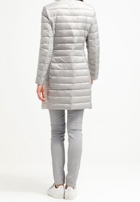 Kobieta w jasnoszarej, pikowanej długiej kurtce, pasujących wąskich spodniach i białych trampkach, stojąca tyłem na jednolitym białym tle.