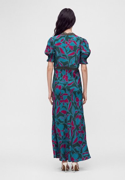 Femme aux longs cheveux foncés portant une robe maxi fleurie turquoise et magenta avec des manches bouffantes et des sandales blanches à talons hauts, vue de dos.
