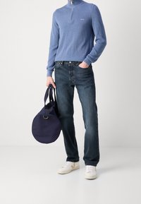 Helles blaues Zip-Hals-Sweatshirt, dunkelblaue Jeans, weiße Sneakers mit beigen Akzenten und eine runde marineblaue Tasche mit Griffen.