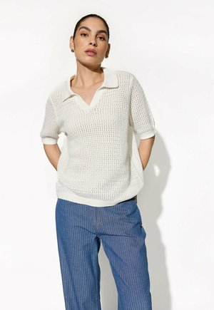 Vrouw met donker haar in een wit, kort mouw gebreid topje en blauwe broek met witte streepjes, poserend tegen een witte achtergrond.