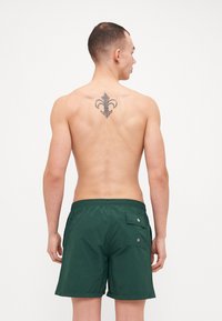 Grønne badebukser med elastisk linning, baklomme og matt finish, båret av en person sett fra bakfra med synlig tatovering.