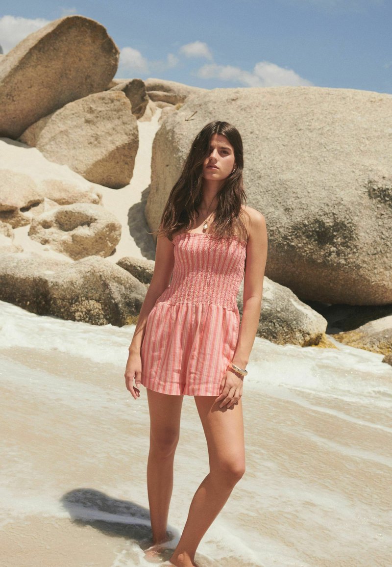 Rosa gestreifter Overall mit gerafftem Oberteil und Shorts, getragen am Strand. Sand und Steine im Hintergrund, mit Wasser am Saum.
