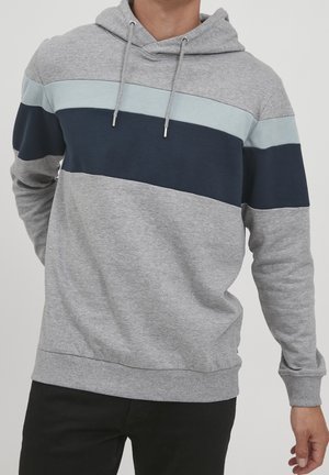 Hombre con sudadera gris con franjas horizontales azul claro y azul marino, de pie contra un fondo liso.