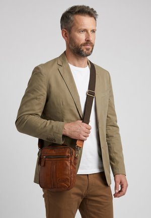 Mann mit kurzen Haaren und Bart trägt einen beigen Blazer und braune Hose, hält eine braune Leder-Umhängetasche mit verstellbarem Riemen.
