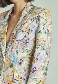 Blazer floreale caratterizzato da un mix di fiori viola chiaro, blu e gialli su uno sfondo crema; con rever a punta e dettaglio della tasca singola.