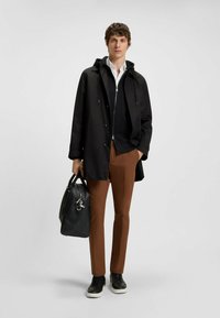 Giacca leggera nera con cappuccio, camicia bianca, maglione nero, pantaloni marroni e sneaker nere. Porta una borsa nera con texture.