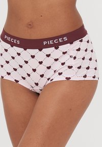 Růžové dámské boxerky s vínovým pasem, na kterém je značka "PIECES". Materiál má vzor srdcí vyznačených přerušovanými čarami.