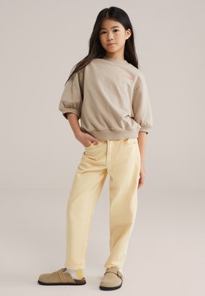 Bež pamučna majica kratkih napuhanih rukava, uparena sa svijetlo žutim trapericama i bež klompama. Jednostavan dizajn bez uzoraka.