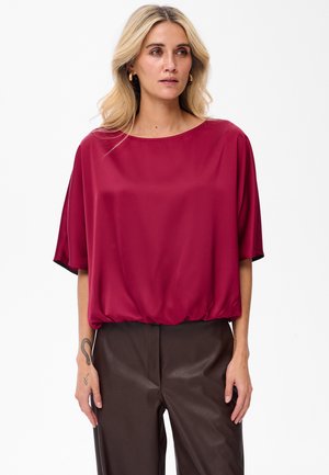 Blonde vrouw met gouden ringoorbellen, een losse donkerroze blouse met driekwartmouwen en donkerbruine broek tegen een witte achtergrond.