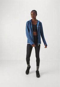 Blå zip-up hoodie med ett texturerat tyg och ett kontrasterande innerfoder, kombinerad med svarta leggings och mörka träningsskor.
