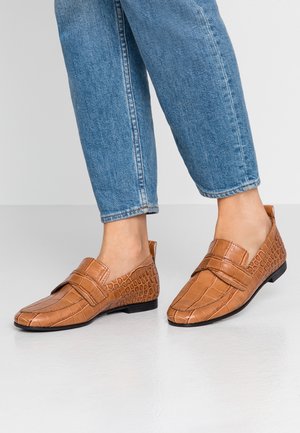 Brune loafers med en krokodillepræget tekstur, en flad sål og en rem over vristen. Båret med blå denimjeans.