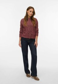 Cable-knit, cropped sweater in rose with button front, drawstring waist, and textured finish. Paired with dark blue flared jeans.