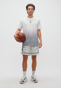 Νεαρός άνδρας ντυμένος με λευκά αθλητικά ρούχα, κρατώντας μια μπάλα μπάσκετ, στέκεται μπροστά σε ένα απλό φωτεινό φόντο.