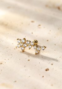 Złota kolczyk wkrętka z kwiatowym wzorem, ozdobiona wieloma przezroczystymi, okrągłymi kryształkami ułożonymi w kwiatowy motyw, na teksturowanym jasnym tle.