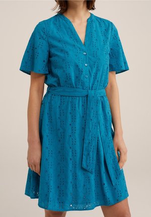 Kobieta ubrana w turkusową sukienkę z krótkim rękawem, zdobioną haftem typu eyelet, z przodu zapinaną na guziki, z dekoltem w serek i pasem z tego samego materiału wiązanym w talii.