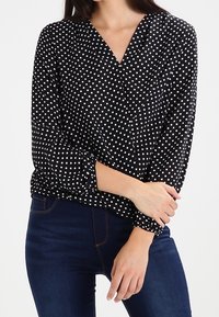 Blusa nera con pois bianchi, scollatura a V, maniche lunghe, polsini arricciati e una texture morbida, abbinata a jeans blu.