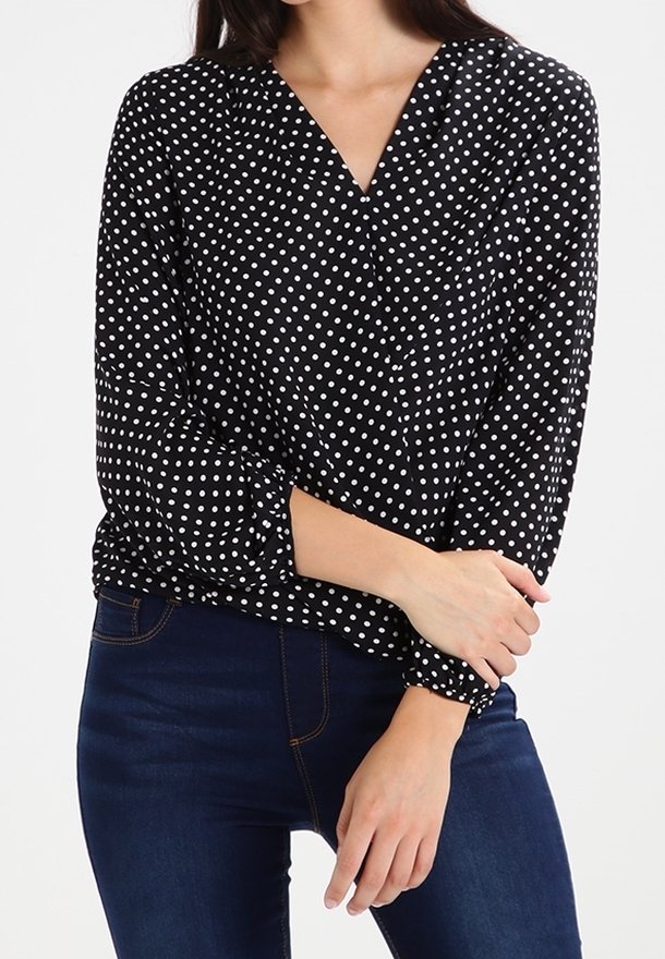 Blusa nera con pois bianchi, scollatura a V, maniche lunghe, polsini arricciati e una texture morbida, abbinata a jeans blu.