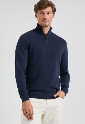 Navy Pullover mit Reißverschlusskragen, gerippten Bündchen und Saum, strukturiertem Strickmuster, getragen über einem hellen Hemd und hellen Hosen.