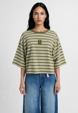 Woman with long dark hair wearing green and white striped oversized T-shirt and loose blue jeans, standing against a plain light background.