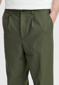 Pantalon vert olive avec une texture lisse, présentant un devant plissé, des poches latérales et une fermeture à bouton.