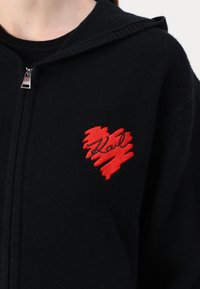 Čierne mikina na zips s červeným srdcovým dizajnom a čiernou výšivkou „Karl“. Materiál pôsobí mäkko a štruktúrovane. S kapucňou.