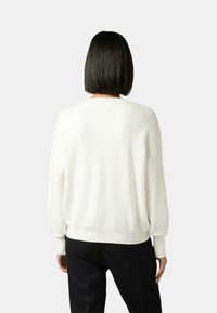 Suéter blanco de punto con un ajuste relajado, hombros caídos, puños acanalados y dobladillo. Llevado con pantalones negros, visto desde la espalda.