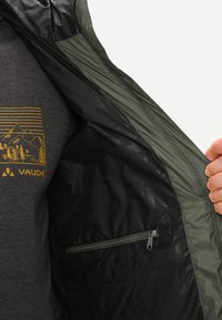 Olijfgroene jas met een zwarte voering, voorzien van een ritszak en een subtiele gemusterde binnenkant. Het materiaal lijkt lichtgewicht en waterafstotend te zijn.